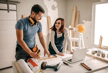 Couple stands in front of a work bench with blueprints on it, smiling and excited to get to work. Construction materials are all around them.
